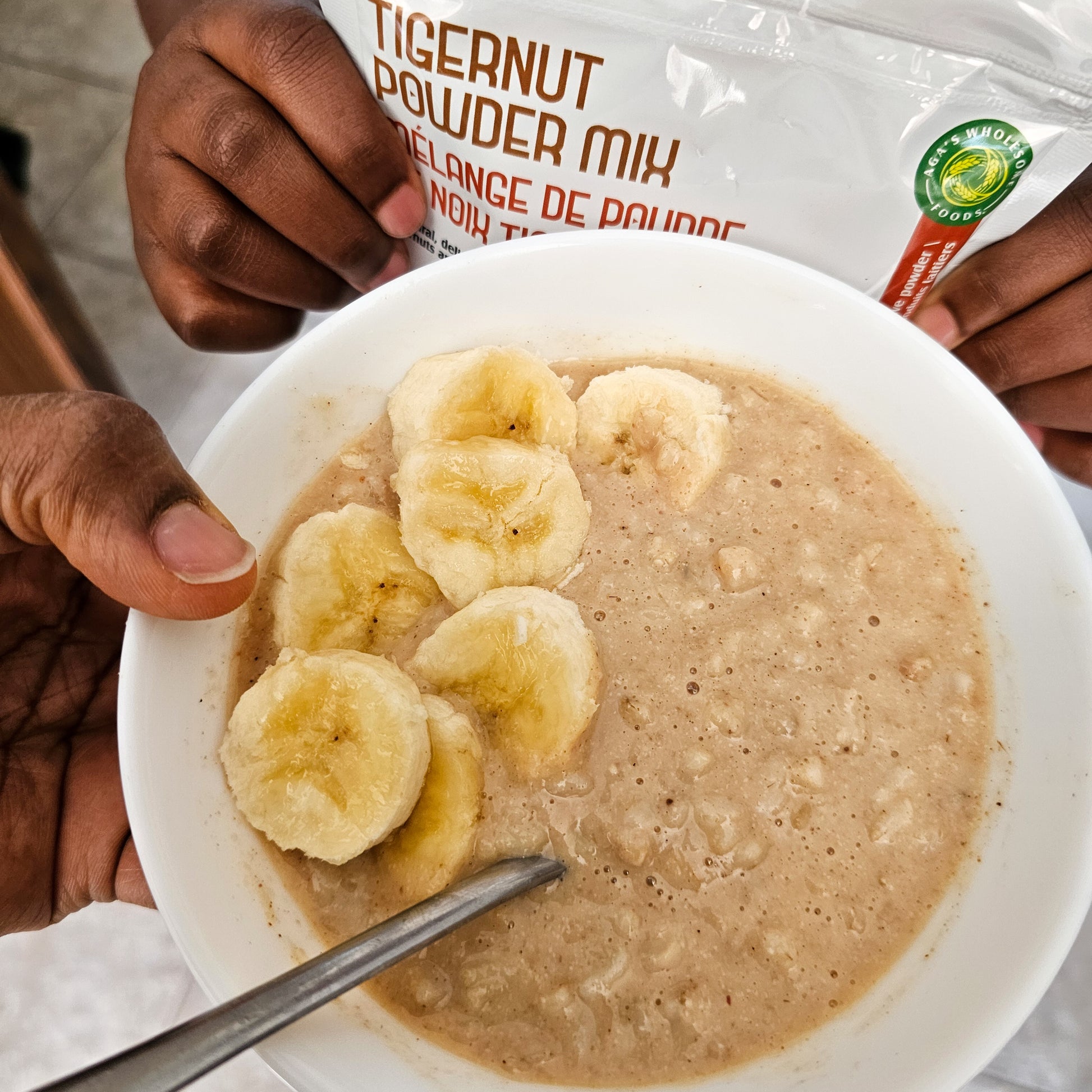 Bowl of tigernut powder mix with sliced bananas and a spoon, with a package of tigernut powder in the background 