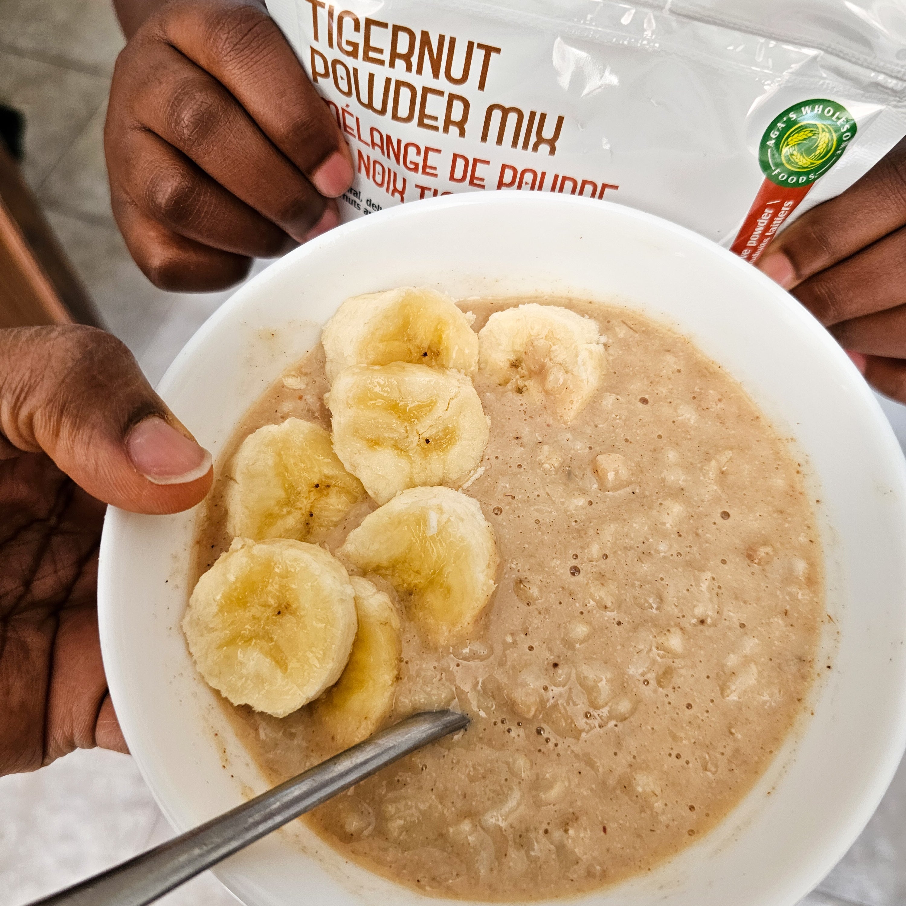 Bowl of tigernut powder mix with sliced bananas and a spoon, with a package of tigernut powder in the background 
